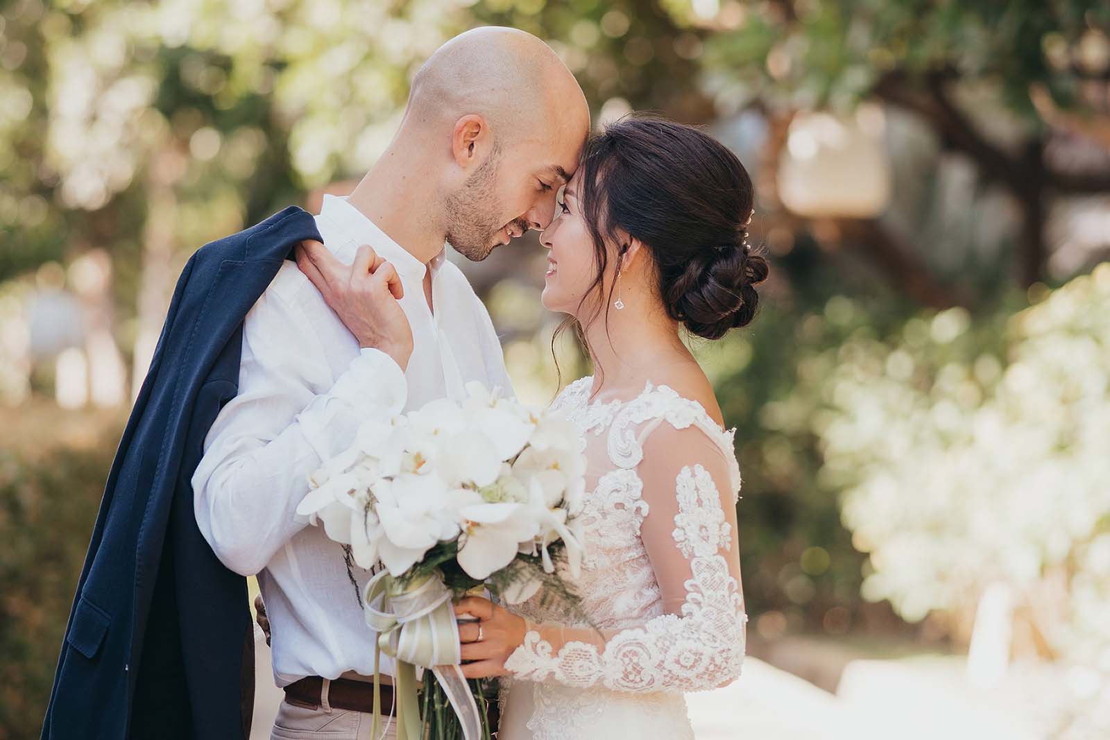 Das Brautpaar steht sich eng gegenüber und blickt sich in die Augen. Die Braut hält einen Brautstrauß aus weißen Orchideen in der Hand.